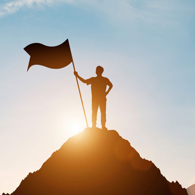 A silhouette of a person holding a flag, on top of a mountain peak, with the sun behind them